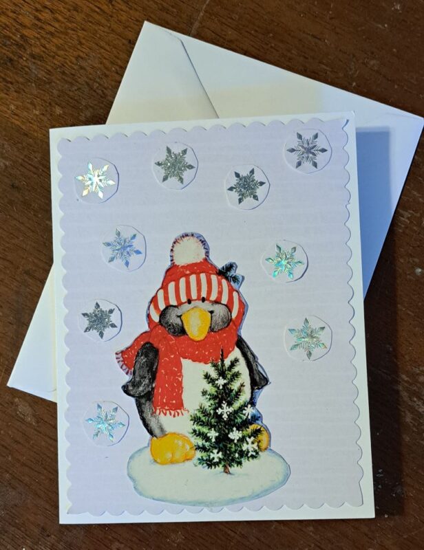 Upcycled Christmas card picturing a penguin wearing a winter hat and scarf standing in front of a tiny pine tree. Surrounding the penguin are cut out snowflakes