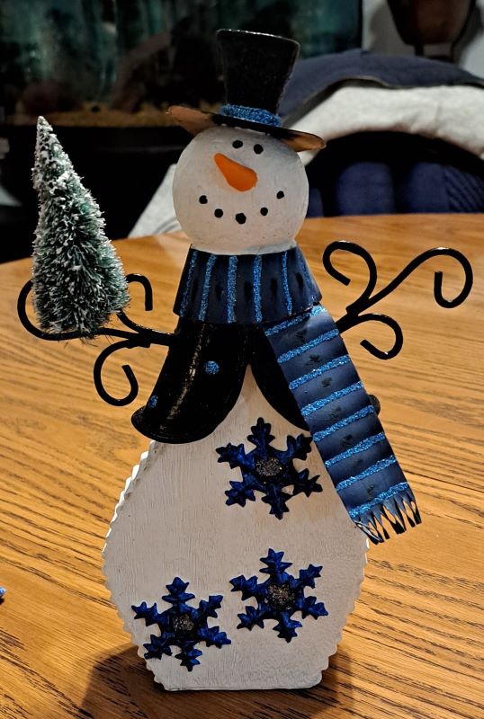 A bright white snow person with black wire arms holding a tiny pine tree. They are wearing a black scarf with light blue glitter stripes, a black vest with blue glitter polka dots. On the body of are blue snowflakes with silver glitter centers.