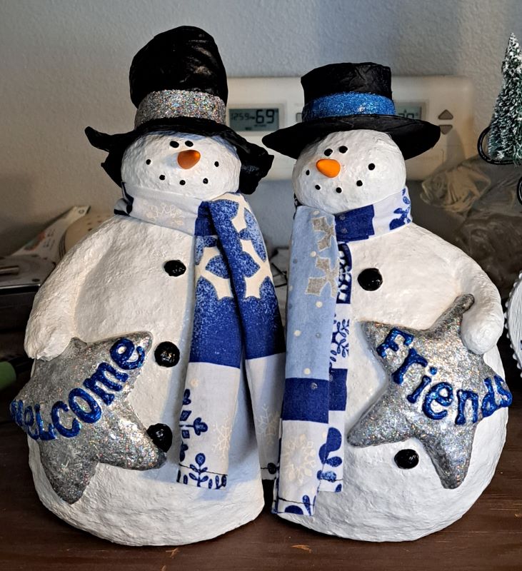 Two bright white snow people with blue and white scarves. They are holding silver stars with blue words that read welcome (left side) and friends (right side). Both are sporting black hats one with silver ribbon and one with blue ribbon.